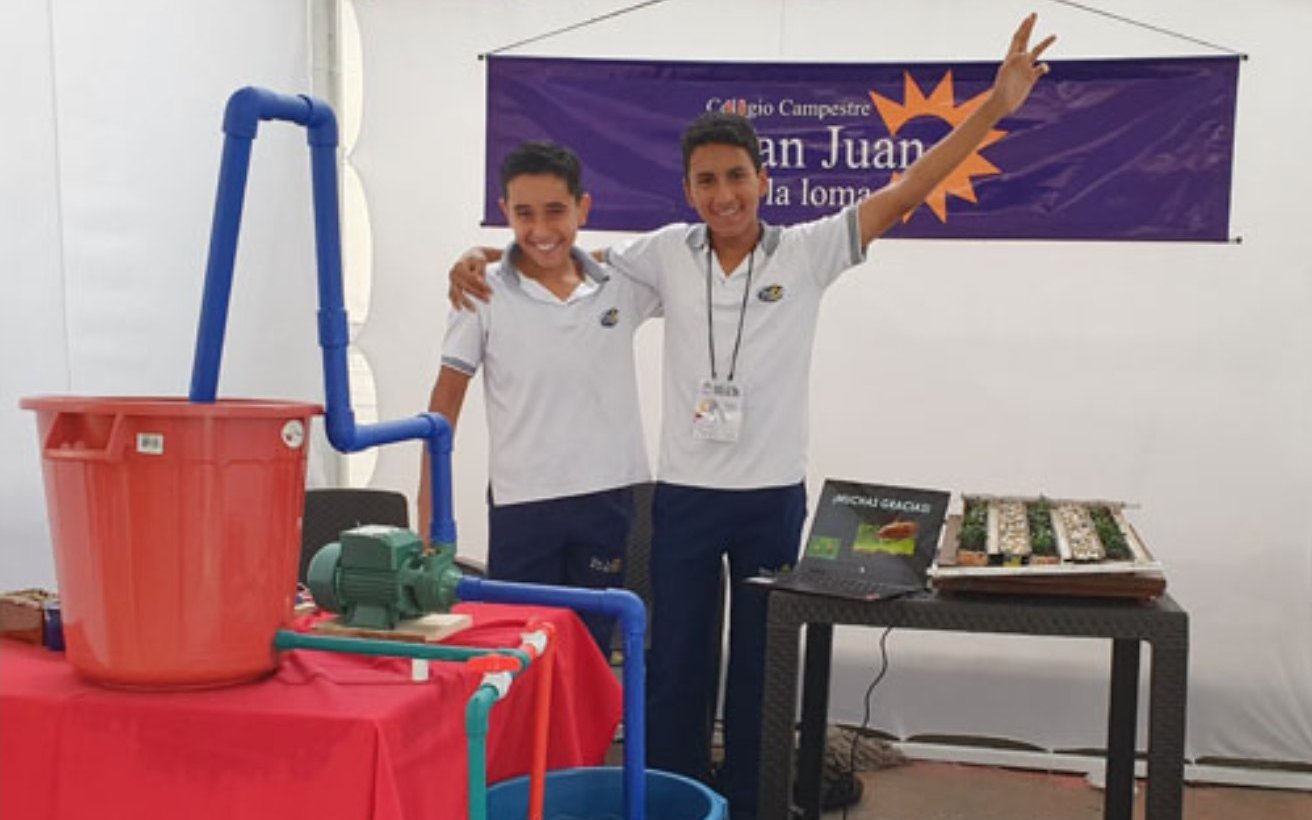 Dos estudiantes con el Bio-Stand: bomba, tubería, tanques y un cultivo con riego automatizado