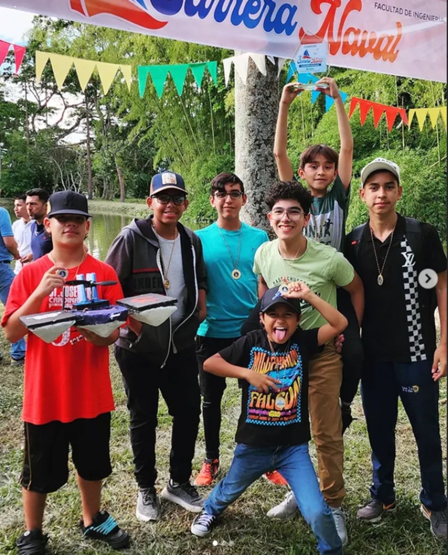 Seis estudiantes del club con medallas y el trofeo bajo la pancarta de la Carrera Naval, uno de ellos con la lancha en las manos