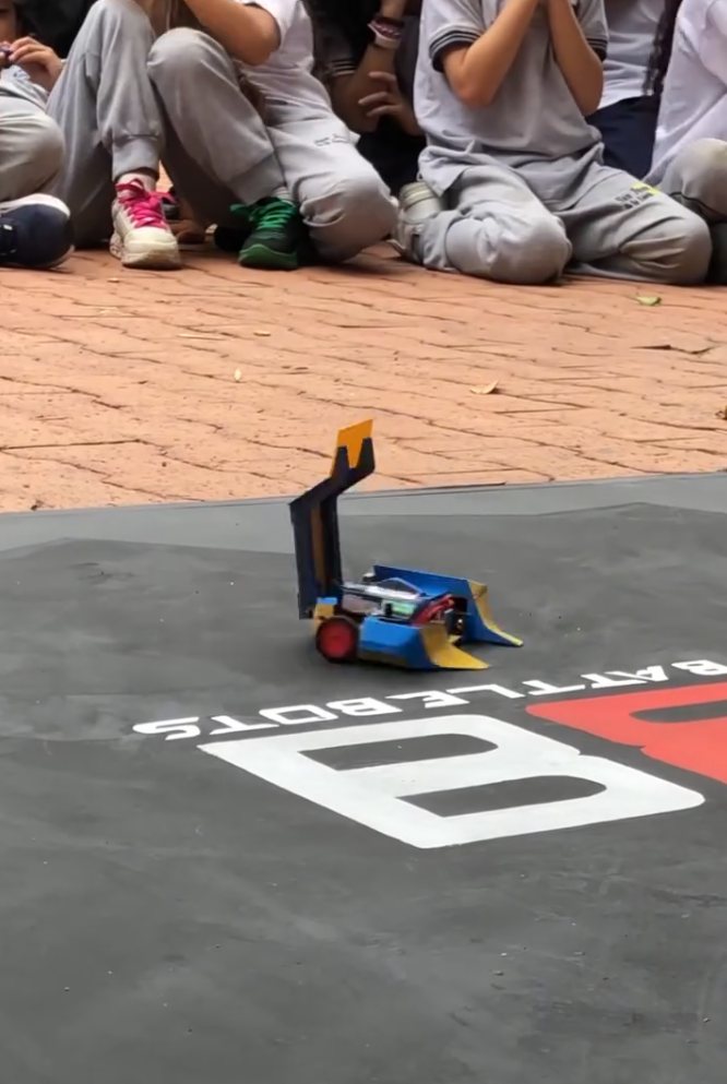 Un robot azul y amarillo con brazo volteador sobre la arena de Robot Battle, con estudiantes sentados alrededor