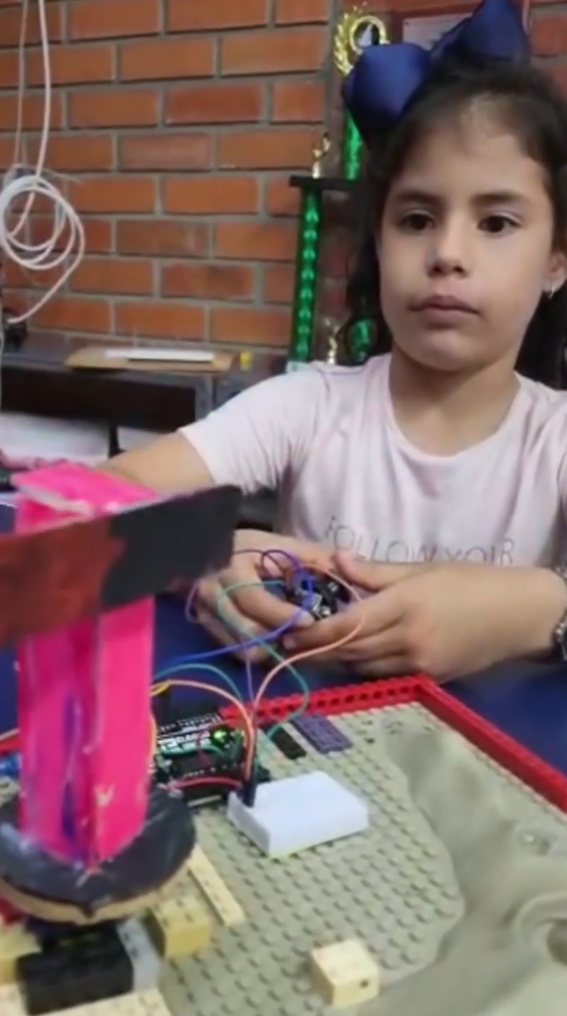 Una estudiante pequeña sostiene los cables de su circuito con Arduino, con una estructura rosada sobre una base de Lego