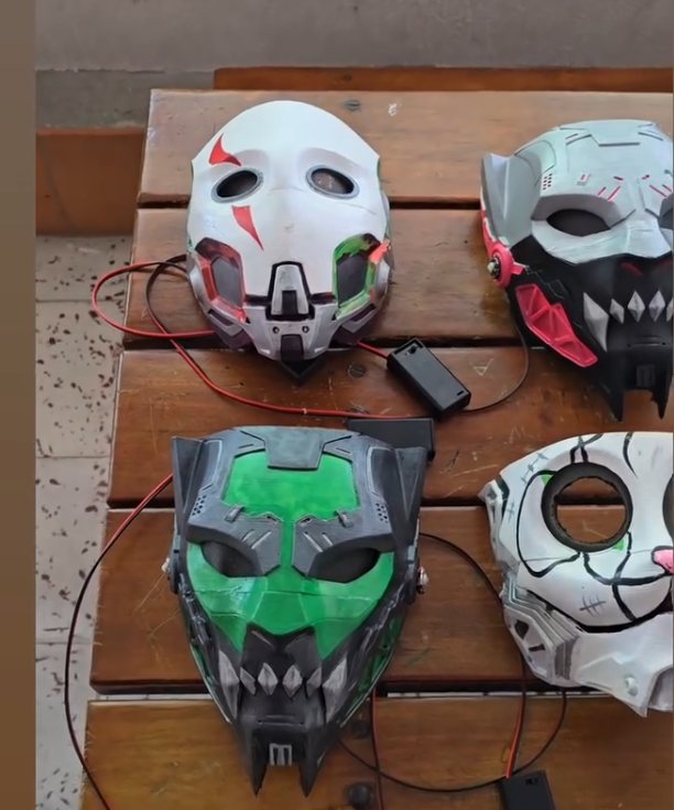 Cuatro máscaras impresas en 3D sobre una mesa de madera: una blanca con detalles verdes, una gris con rojo, una verde con dientes y una calavera blanca